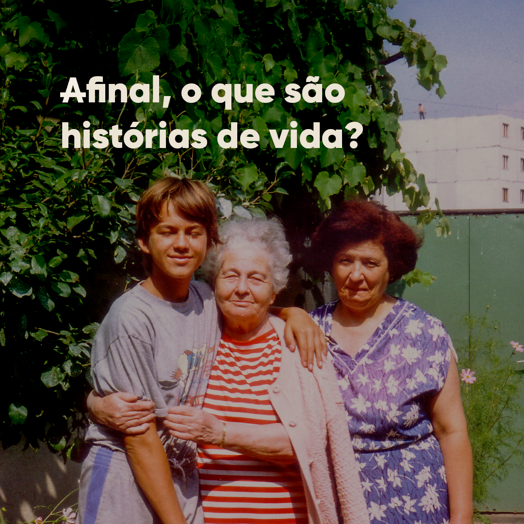 Um menino é abraçado por uma senhora de cabelos brancos e blusa listrada vermelha. Ao lado dela, tem outra senhora, de vestido florido azul e cabelos castanhos curtos. Atrás dos três tem uma árvore.